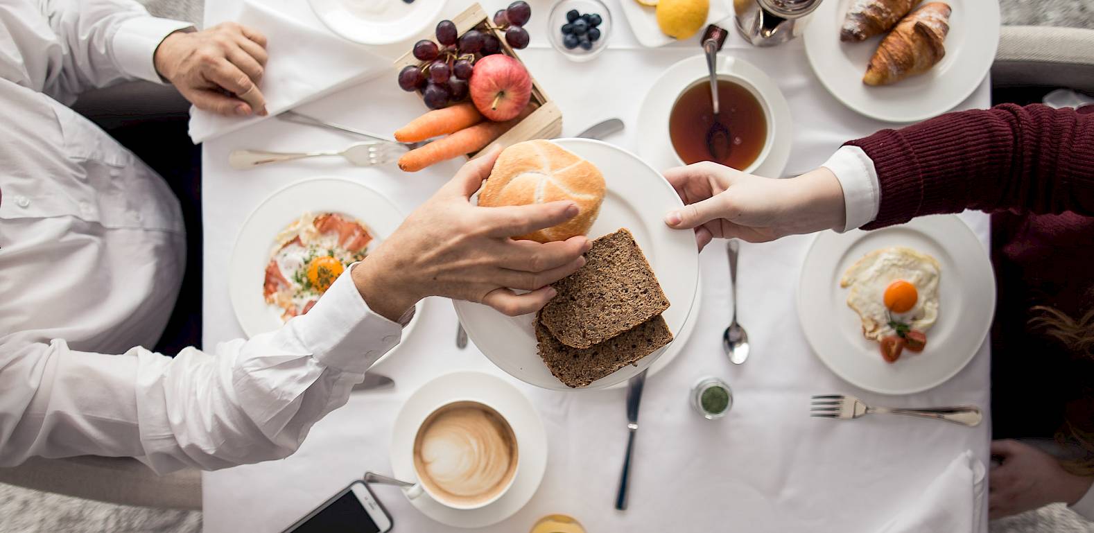 Frühstücksszene mit verschiedenen Lebensmitteln auf einem Tisch. Hände reichen sich ein Brot.