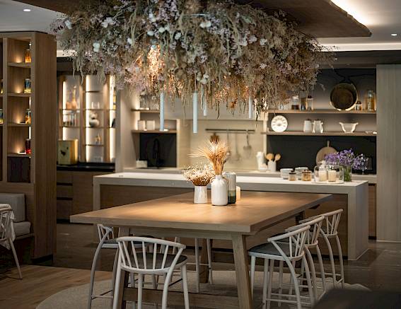 Moderne Küche mit Holz-Esstisch, weißen Stühlen und einem hängenden Blumenarrangement von der Decke.