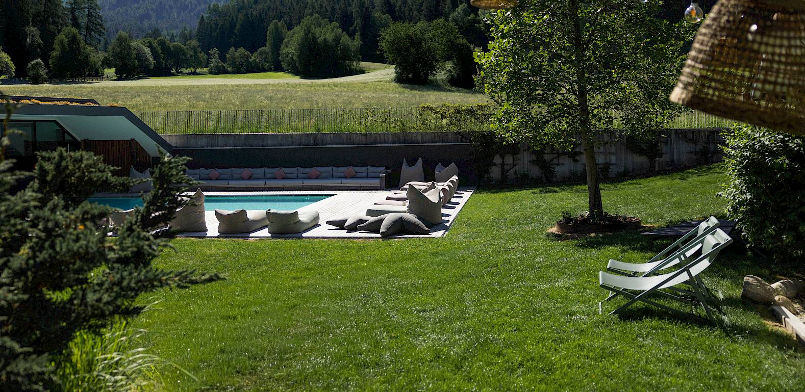Grüne Wiese mit Liegestühlen, Pool im Hintergrund, umgeben von Bäumen und Bergen unter freiem Himmel.