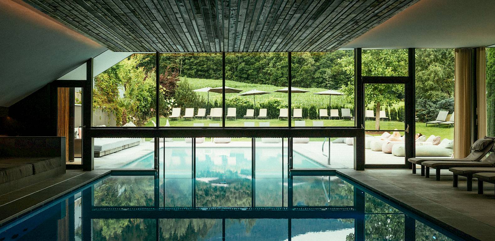 Innenpool mit großen Glasfenstern, Blick auf Liegestühle im Garten. Holzdecke und natürliche Beleuchtung.