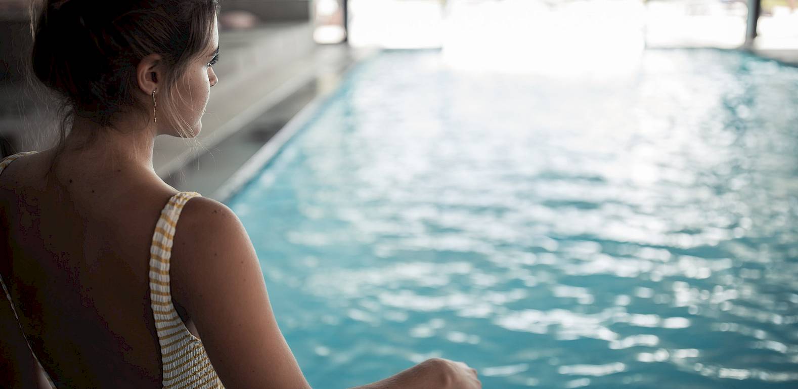 Frau in gestreiftem Badeanzug schaut auf ein Hallenbecken. Natürliches Licht beleuchtet das Wasser.