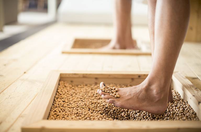 Barfuß gehen zwei Füße über kleine Erbsen in einem Holzkasten für eine Fußmassage.