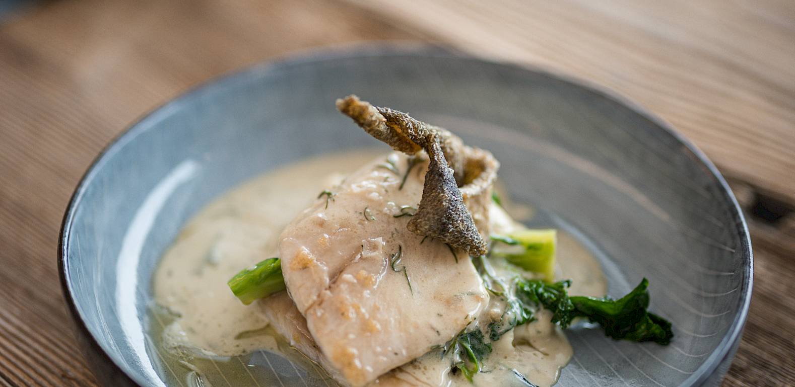 Gegrillter Fisch auf Spinatbett mit cremiger Sauce in einer grauen Schale auf Holztisch.