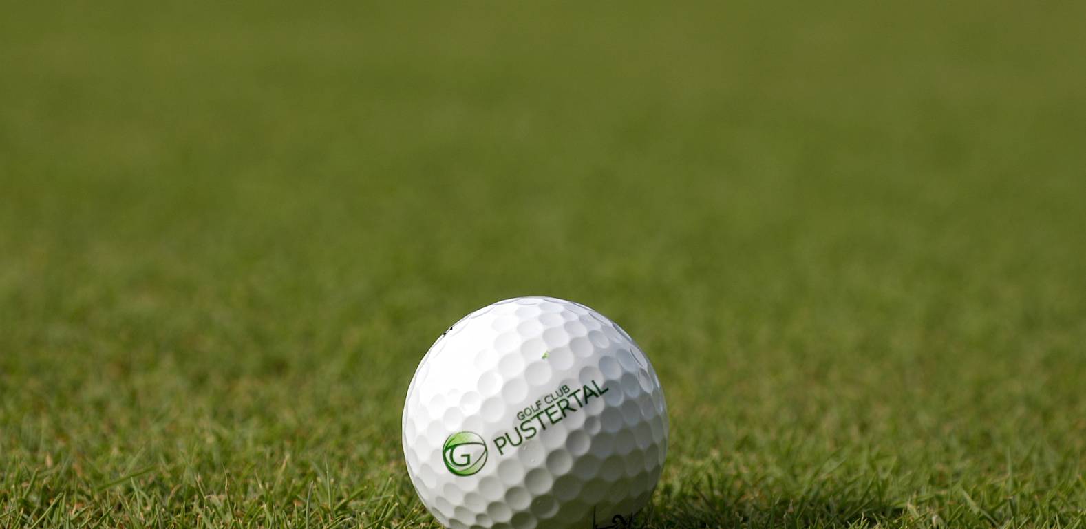 A golf ball with the "G Pustertal" logo rests on well-maintained grass.