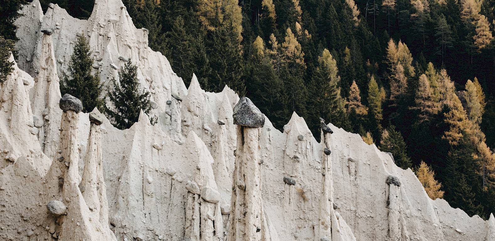 Steinerne Erdpyramiden vor einem herbstlichen Wald mit gelbem und grünem Laub.