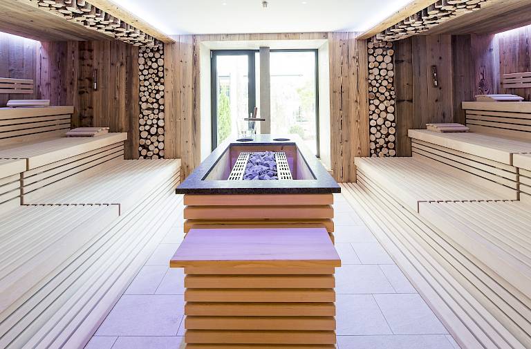 Eine moderne Sauna mit Holzbänken und zentralem Ofen, warme, helle Beleuchtung. Fenster im Hintergrund.