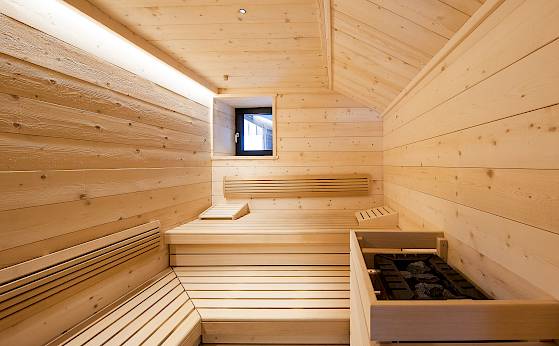 Holzsauna mit Bänken und Ofen, kleines Fenster an der Wand, warme Beleuchtung. Entspanntes Ambiente.