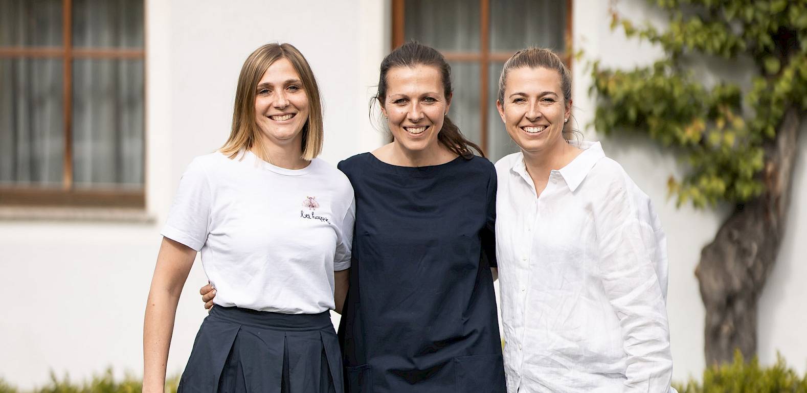 Tre donne sorridenti in piedi insieme davanti a un edificio bianco con finestre e piante sullo sfondo.
