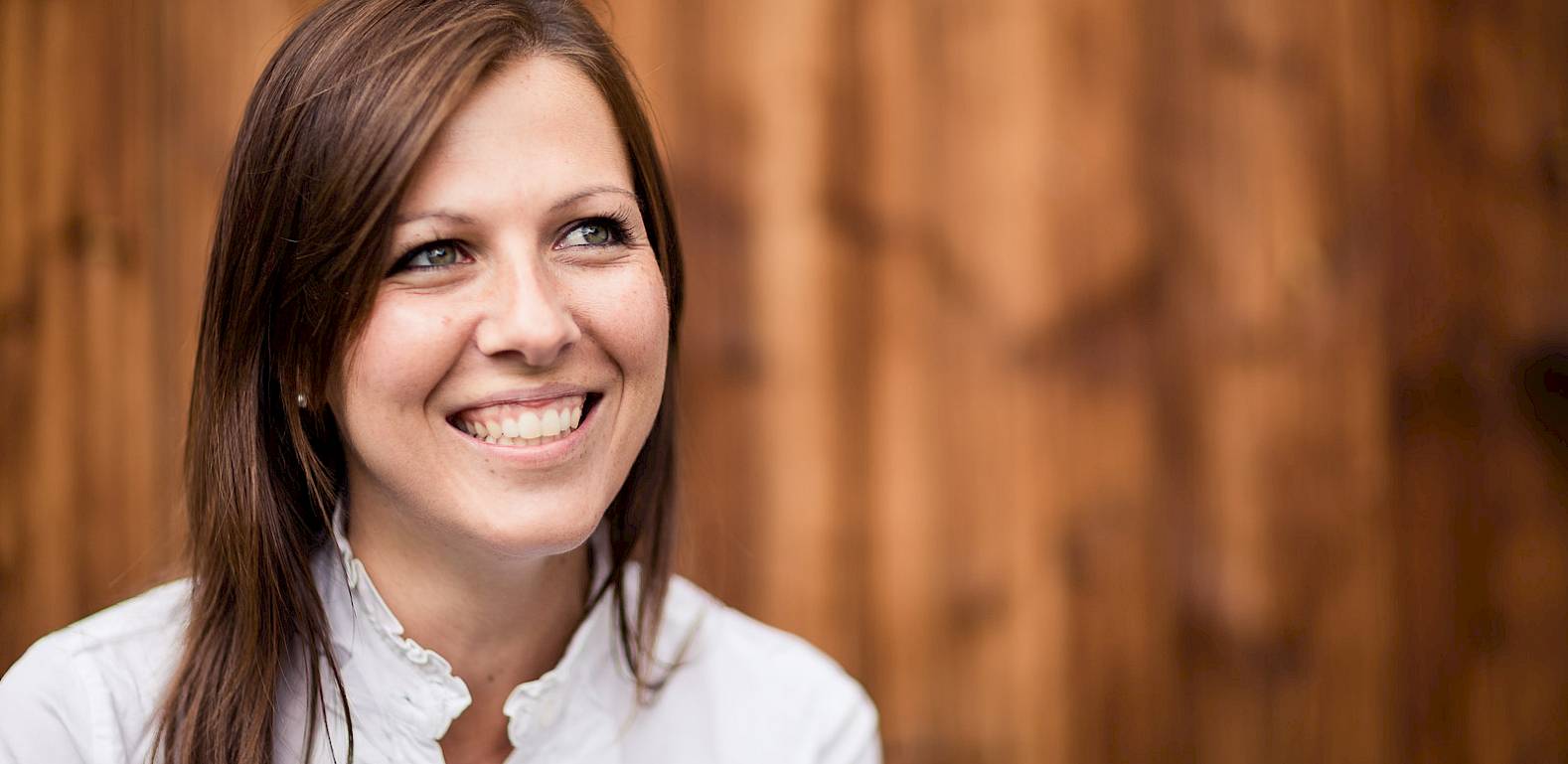 Una donna sorridente in camicetta bianca davanti a uno sfondo di legno sfocato.