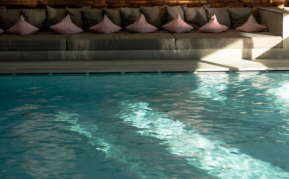 Ein Hallenbad mit türkisfarbenem Wasser, Holzwand und einer Sitzreihe mit rosa Kissen im Hintergrund.