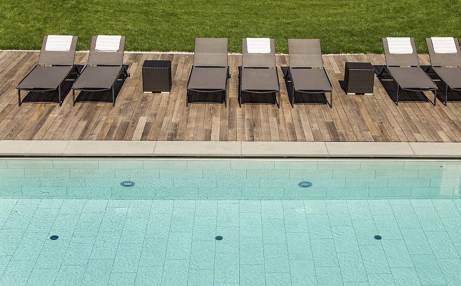 Liegestühle auf einer Holzterrasse am Pool, ein grüner Rasen im Hintergrund.