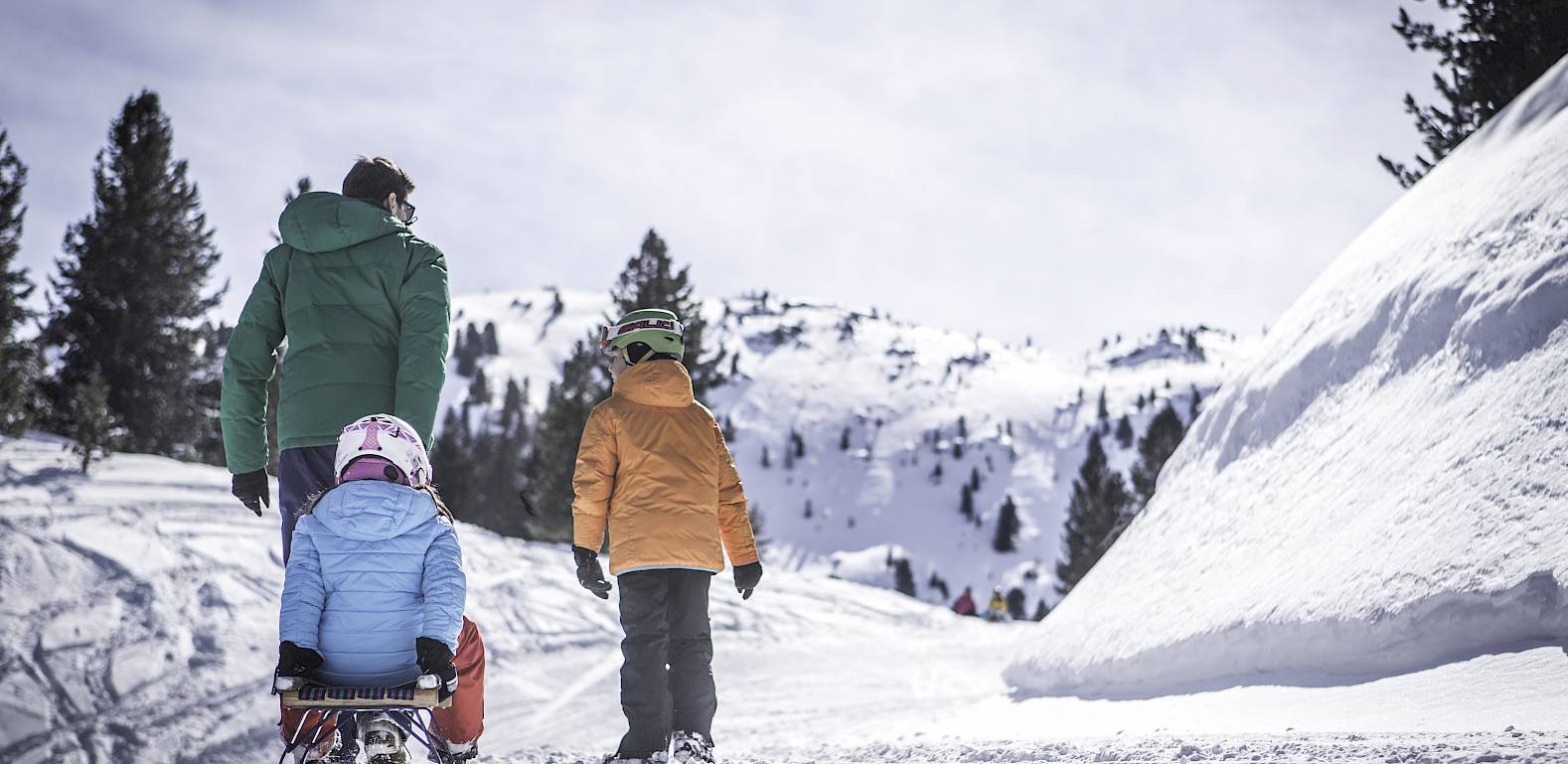 Erwachsener und zwei Kinder im Schnee beim Schlittenfahren, umgeben von verschneiten Bergen und Bäumen.