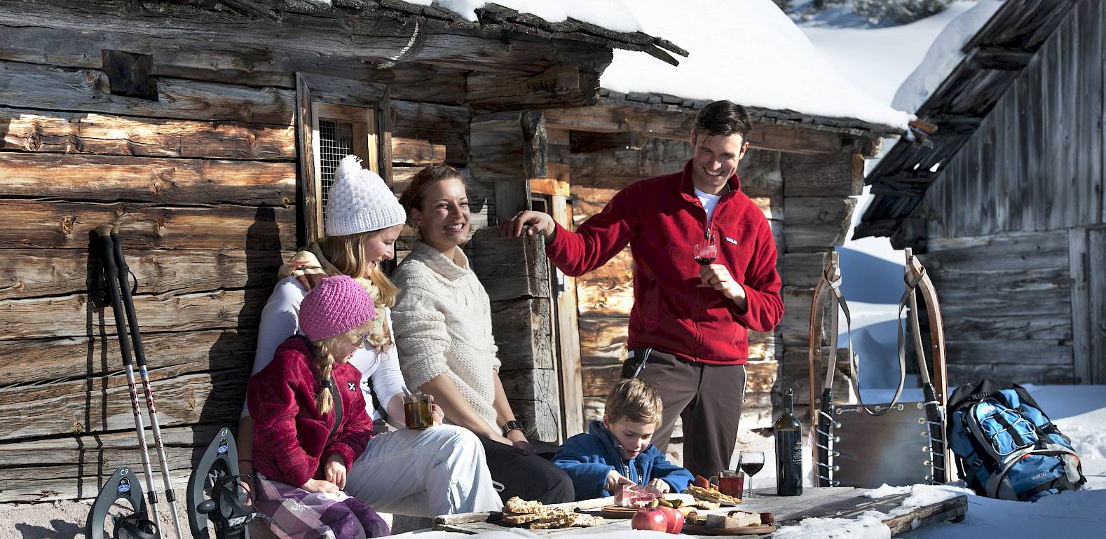 Familie genießt ein Picknick im Schnee vor einer Holzhütte, mit Bergen im Hintergrund. Wintersportausrüstung sichtbar.