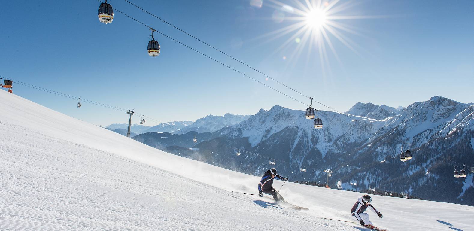 Zwei Skifahrer fahren eine schneebedeckte Piste hinab, während die Sonne über den Bergen strahlt.