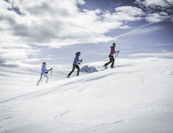 Drei Personen beim Skitourengehen auf einem verschneiten Hang, unter teils bewölktem Himmel.