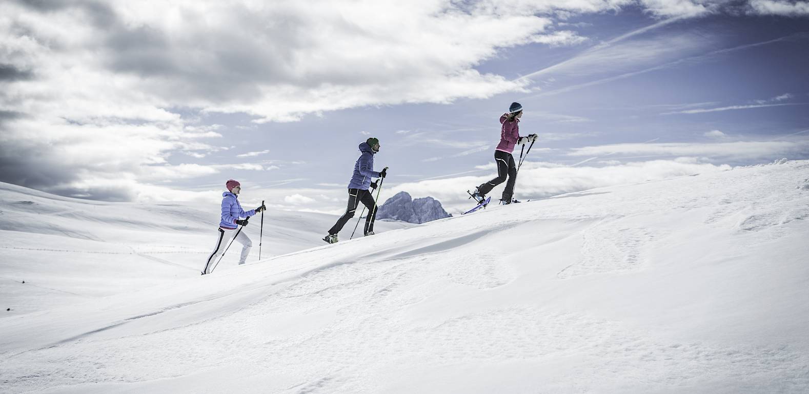 Drei Personen beim Skitourengehen auf einem verschneiten Berghang unter bewölktem Himmel.