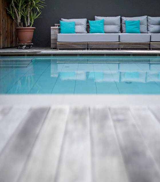 Holzterrasse, Pool und graue Sofas mit türkisfarbenen Kissen, dekoriert mit grüner Pflanze.