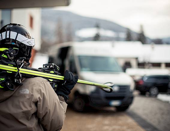 Person in Skiausrüstung hält Skier vor einem Transporter, verschneite Landschaft im Hintergrund.