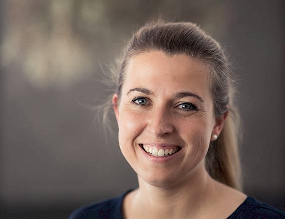 A smiling woman with brown hair in a dark top against a blurred background.