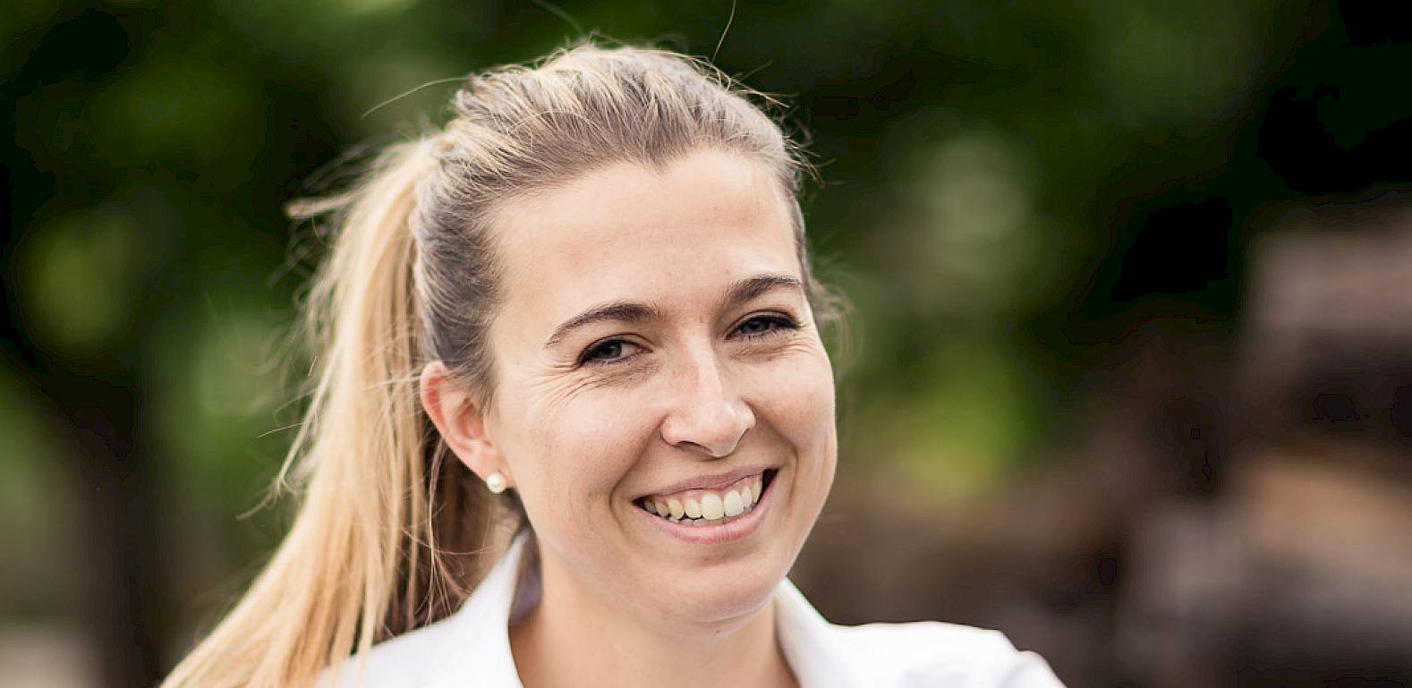 Una donna sorridente con camicia bianca all'aperto. Sfondo verde sfocato.