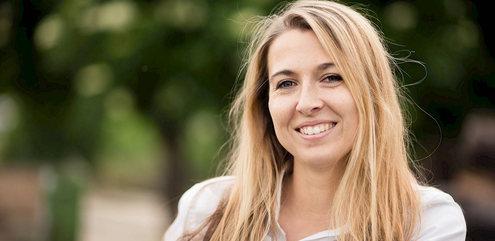 Una donna bionda sorridente all'aperto davanti a uno sfondo verde sfocato.