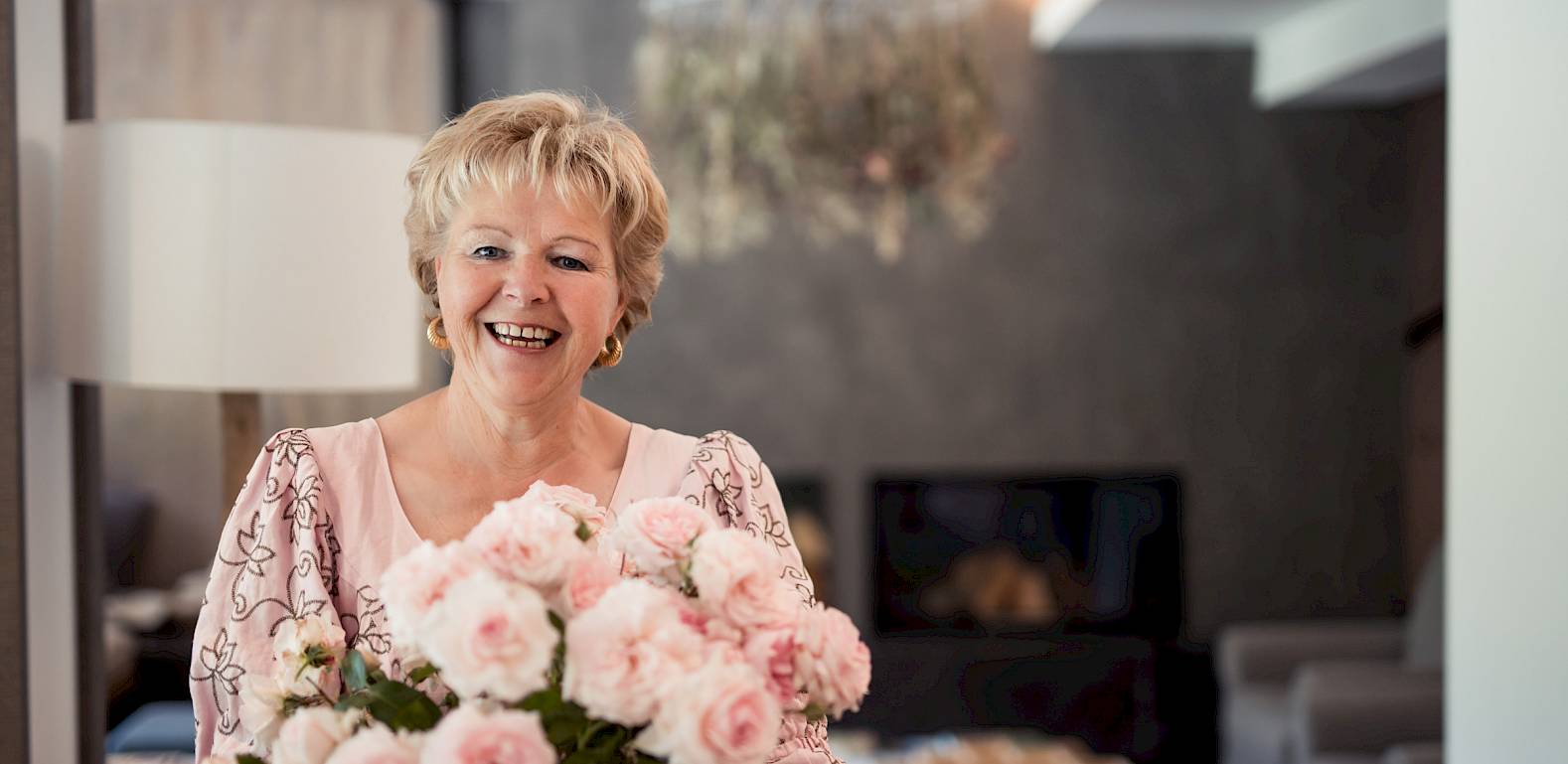Anziana donna sorride accanto a un mazzo di rose rosa in un soggiorno elegantemente arredato.
