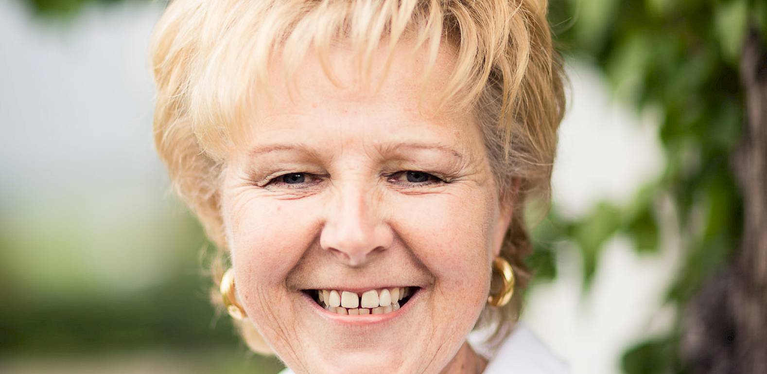 Una donna sorridente con capelli corti biondi e camicia bianca davanti a un albero nel verde.