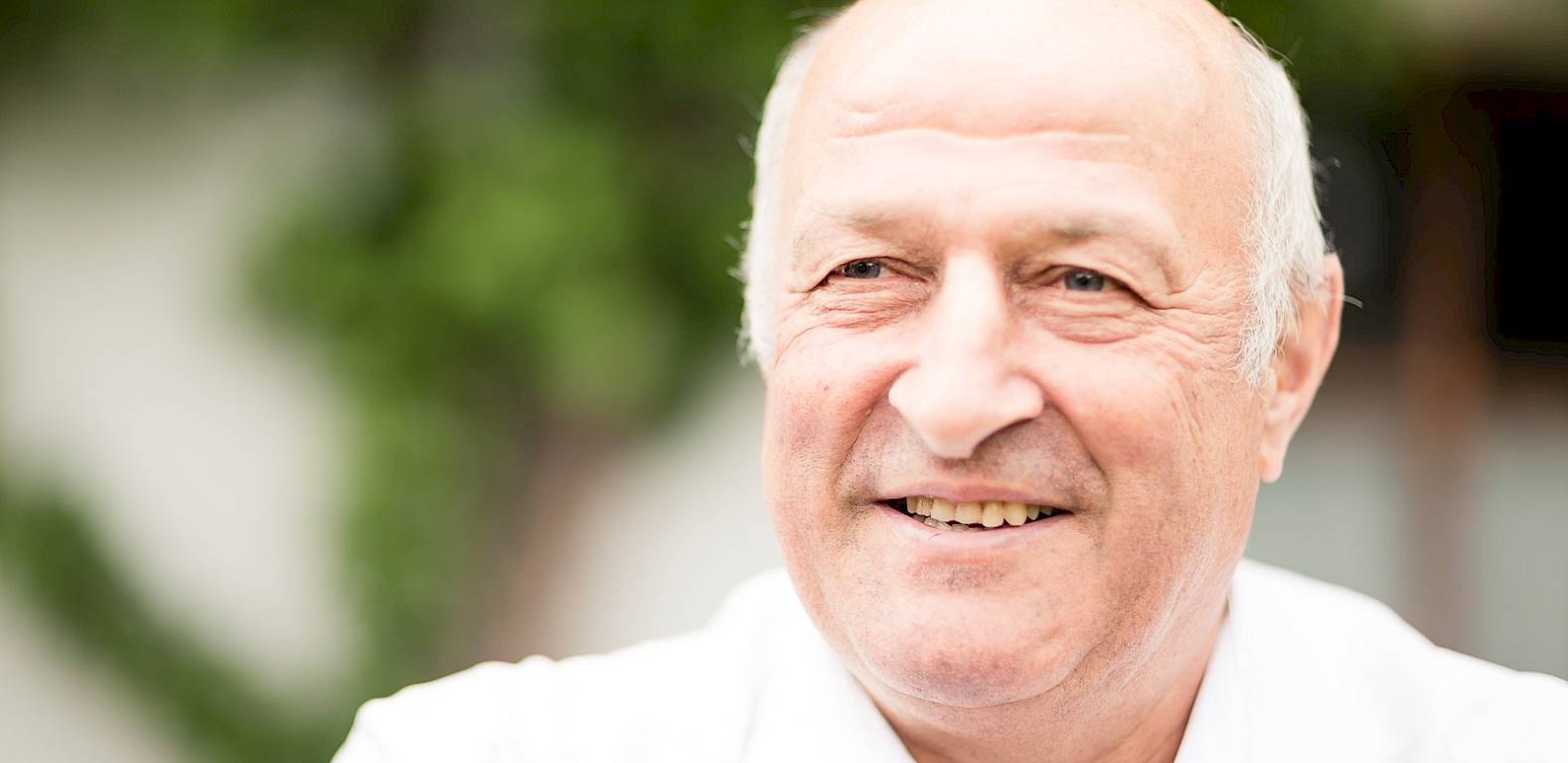 Un uomo anziano calvo sorride all'aperto. Sfondo verde sfocato.