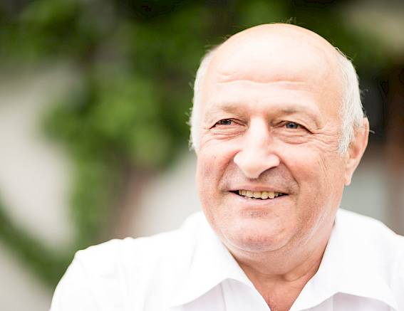 Ein älterer Mann lächelt freundlich in einem Garten. Er trägt ein weißes Hemd.
