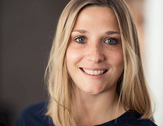 Una donna sorridente con capelli biondi indossa un top scuro e guarda verso la fotocamera.