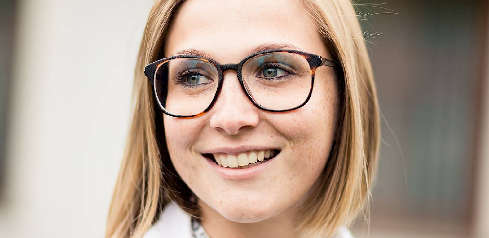 Una donna sorridente con occhiali e camicetta bianca davanti a uno sfondo sfocato all'aperto.