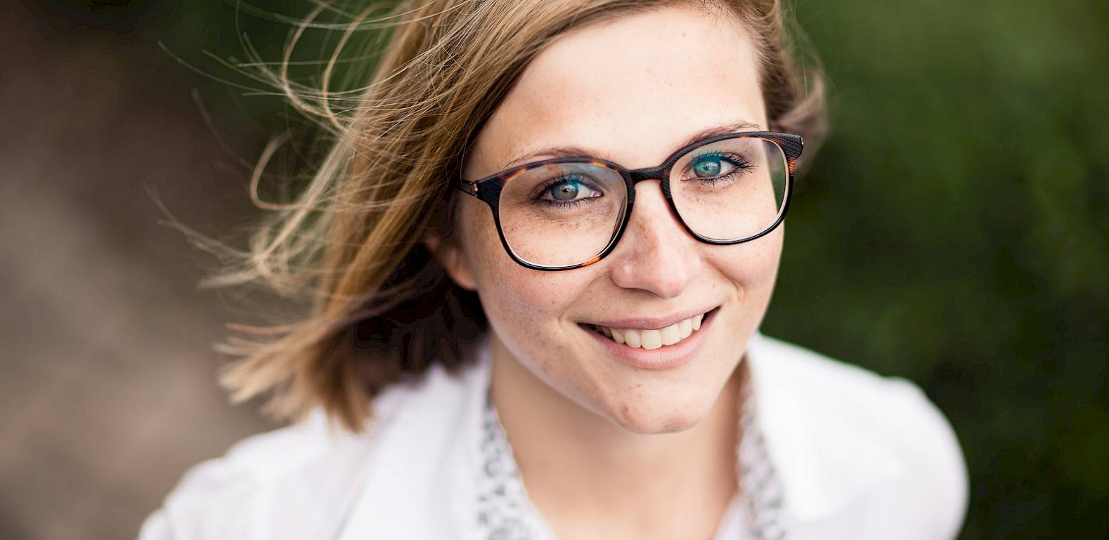 Donna con occhiali e sorriso, indossa una camicia bianca. Sfondo sfocato.