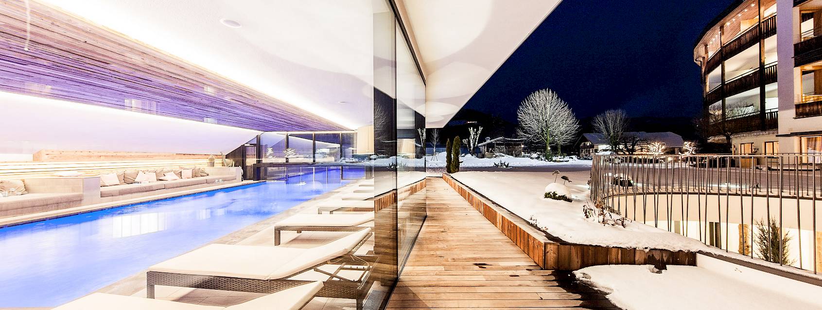 Beheizter Pool bei Nacht neben einer Terrasse mit Liegestühlen, umgeben von schneebedeckter Landschaft.