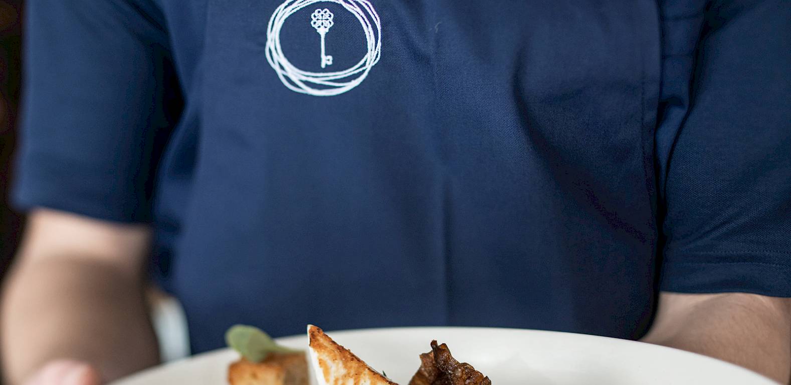 Un cameriere con grembiule blu scuro serve un elegante piatto con pane tostato e funghi su un piatto bianco.