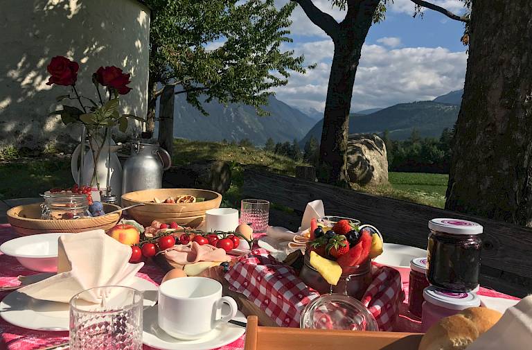 Colazione all'aperto con vista sulle montagne, la luce del sole illumina una tavola ricca di cibo.