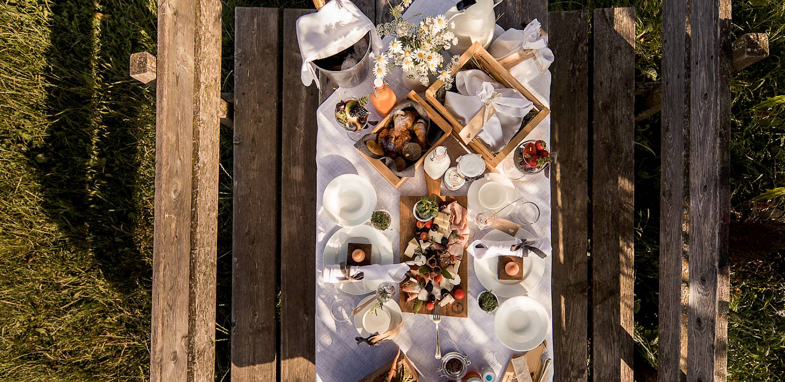 Picknicktisch im Grünen, gedeckt mit verschiedenen Speisen und Getränken auf weißem Tuch. Luftaufnahme.