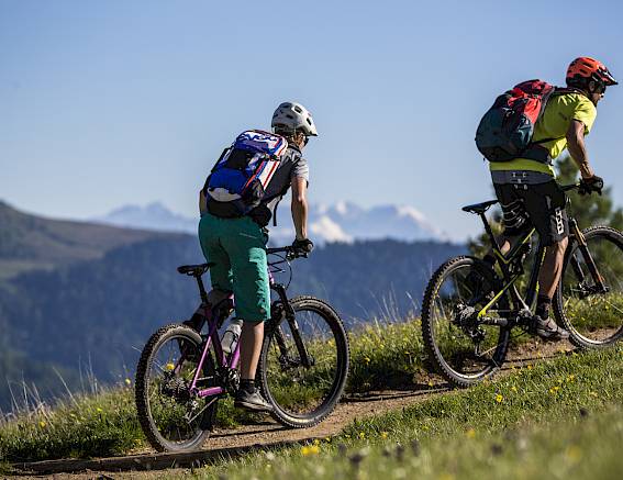 Zwei Radfahrer fahren einen Hügelpfad hinauf, im Hintergrund Berge und blauer Himmel.