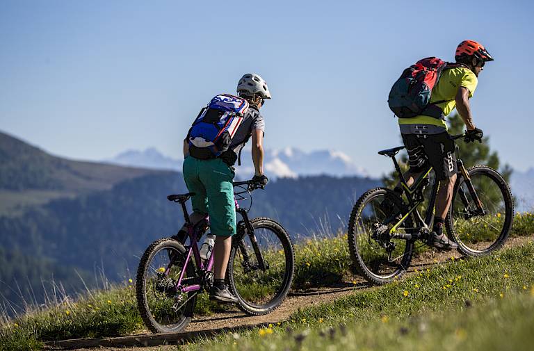 Due ciclisti salgono un sentiero collinare, con montagne e cielo blu sullo sfondo.