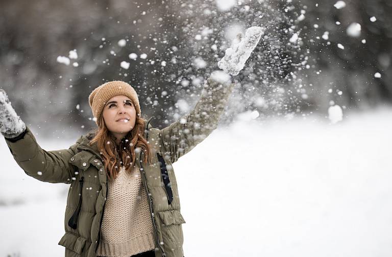 Donna con cappotto invernale e cappello lancia neve in aria, circondata da un paesaggio innevato.