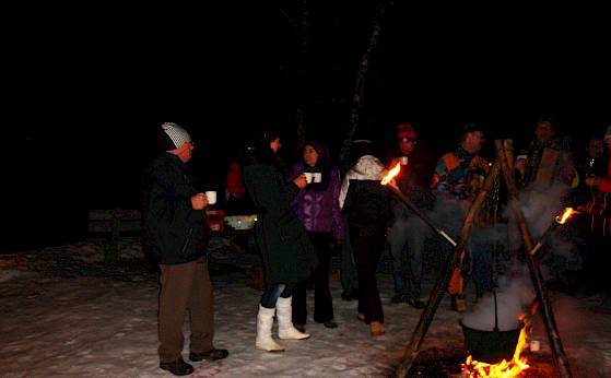 People gather around a campfire in the snow. Some hold cups, enjoying the warmth.