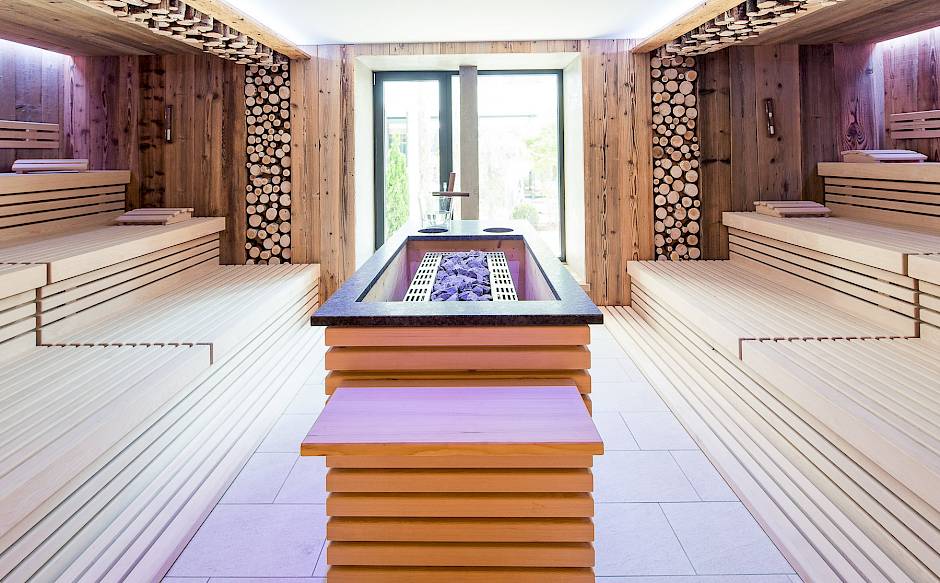 Spacious modern sauna room with wooden benches and central stove, naturally lit by glass windows.