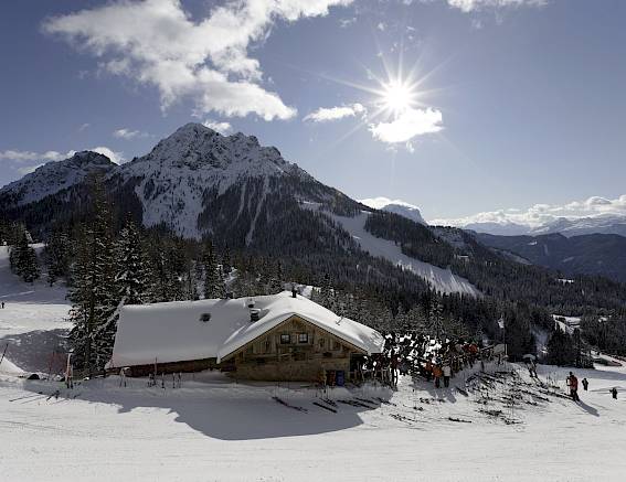 Verschneite Berghütte mit Bergen im Hintergrund unter sonnigem Himmel. Menschen sind auf einer Skipiste zu sehen.
