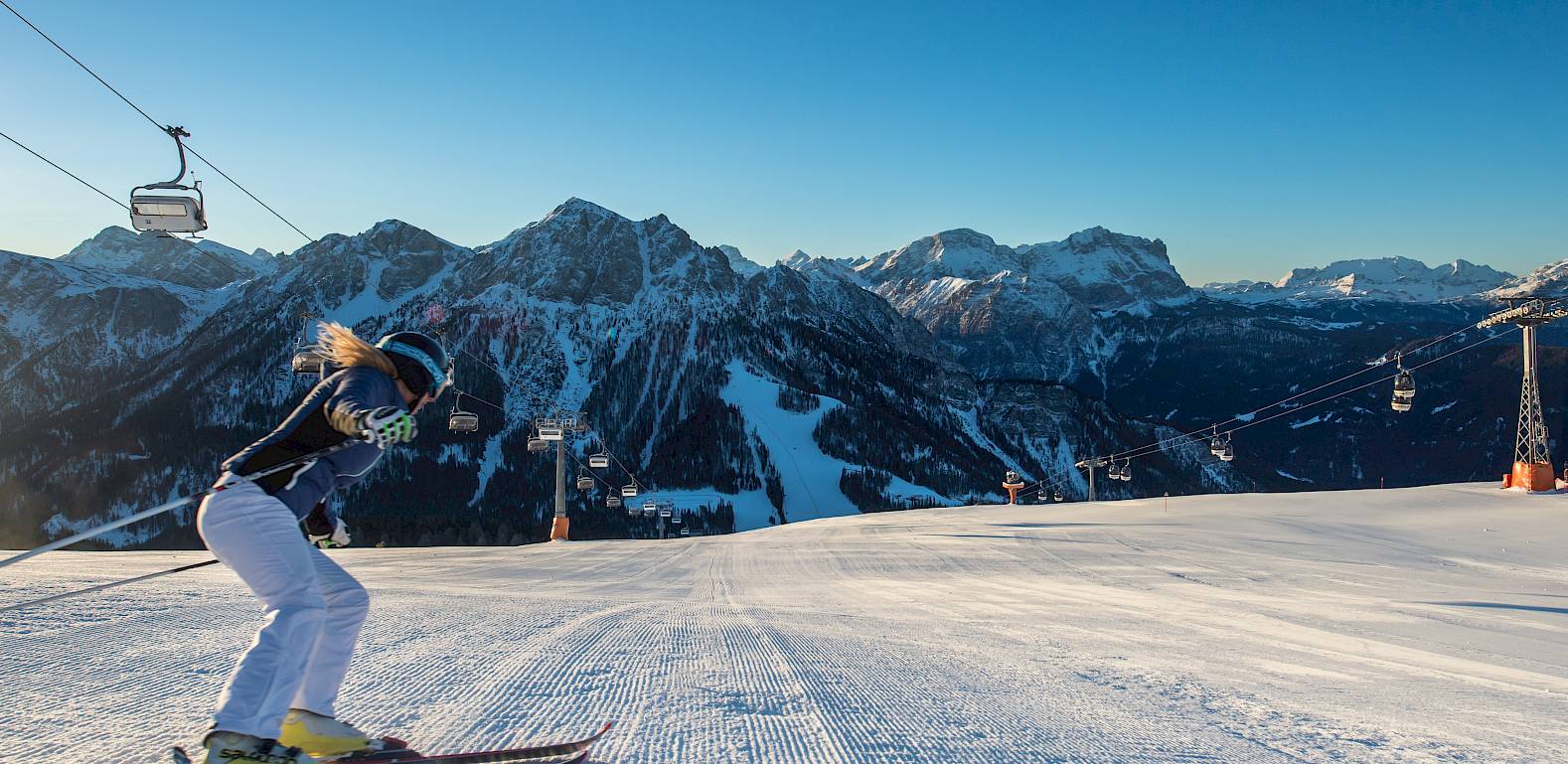 Eine Skifahrerin fährt eine frisch präparierte Piste hinunter, umgeben von verschneiten Bergen unter klarem Himmel.