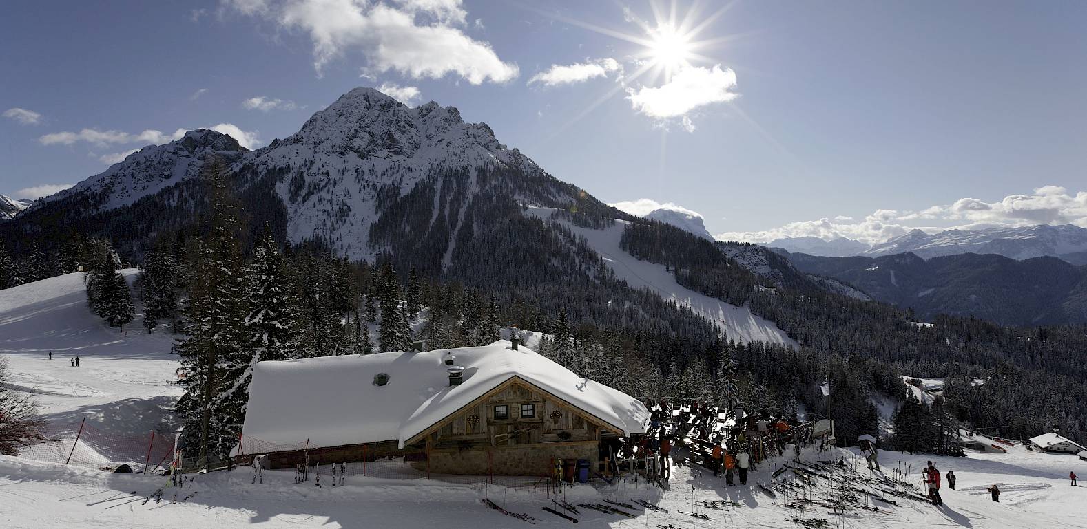 Berglandschaft mit Hütte im Vordergrund, schneebedeckte Berge und strahlender Sonnenschein.
