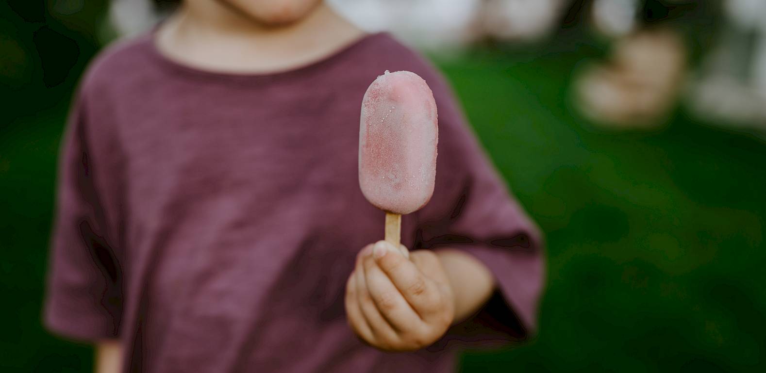 Bambino in maglia viola tiene un gelato rosa su prato verde, lingua fuori.
