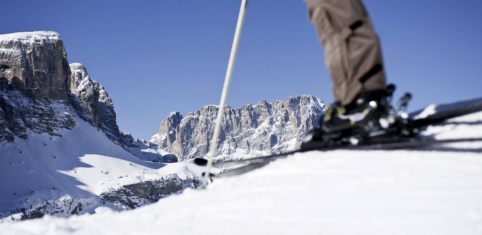 Sciatore sul bordo di una pista con montagne innevate sullo sfondo sotto un cielo blu chiaro.