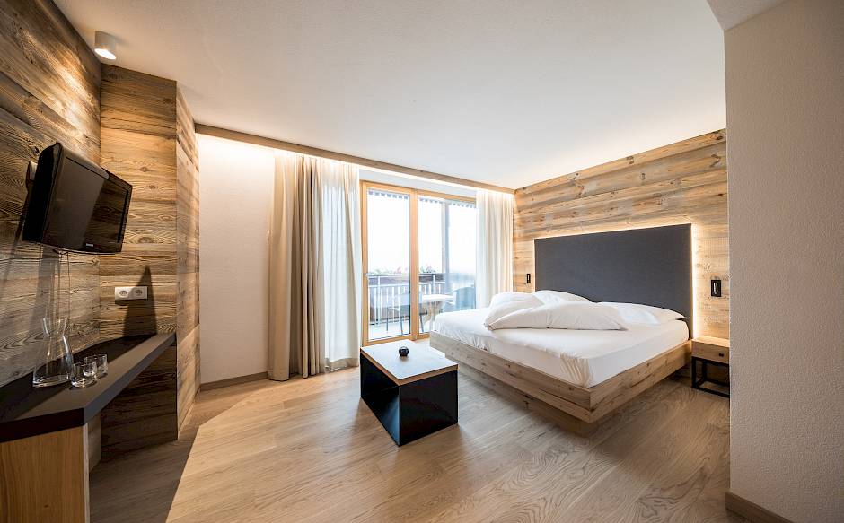 Camera d'albergo moderna in legno con letto matrimoniale, TV e grande finestra che lascia entrare molta luce.