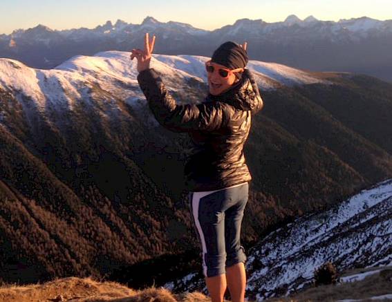Person on mountain peak making peace sign, surrounded by snow-covered mountains under clear sky.