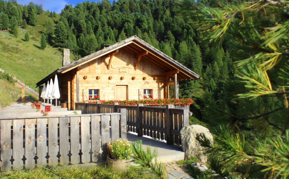 Holzhaus in einer alpinen Landschaft, umgeben von grünen Bäumen und einem blauen Himmel im Hintergrund.