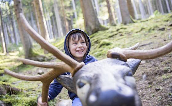 Ein lachendes Kind mit blauer Kapuze sitzt auf einer Holzskulptur eines Rentiers im Wald.
